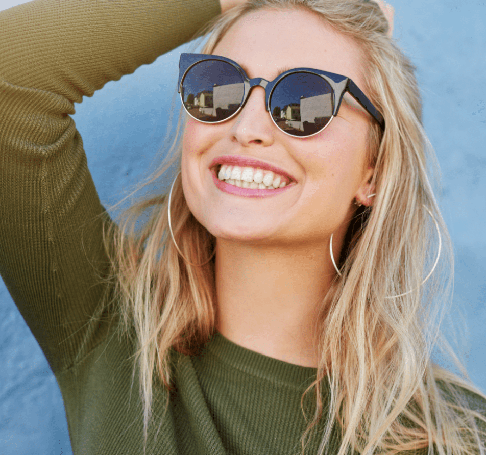 Young woman wearing a green shirt smiling wearing sunglasses in front of a blue background