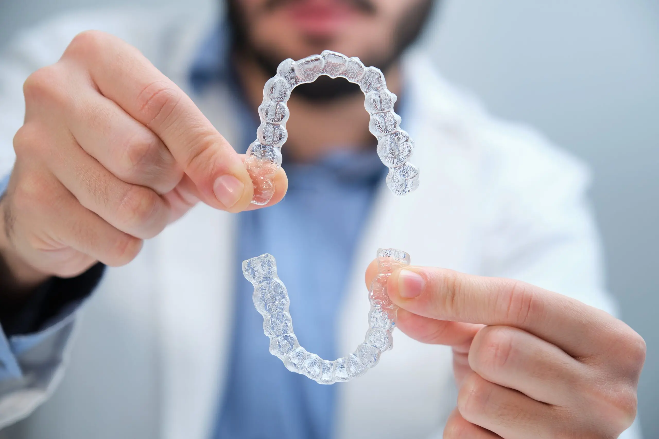 A person in a white coat holds two clear plastic retainers, one in each hand, with their face out of focus in the background.