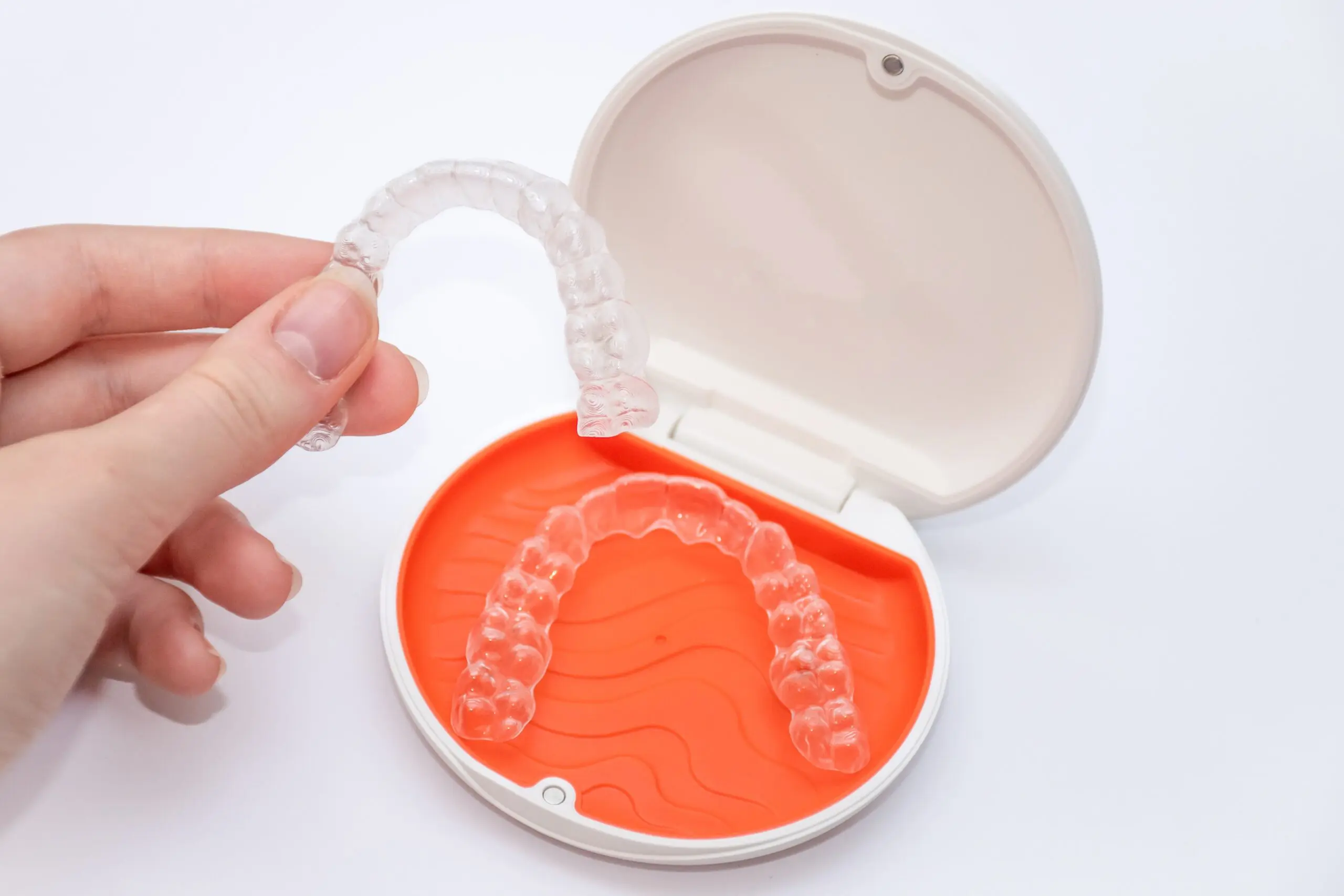 A hand holds a clear plastic retainer above an orange-lined case with another aligner inside, both designed for orthodontic teeth straightening.