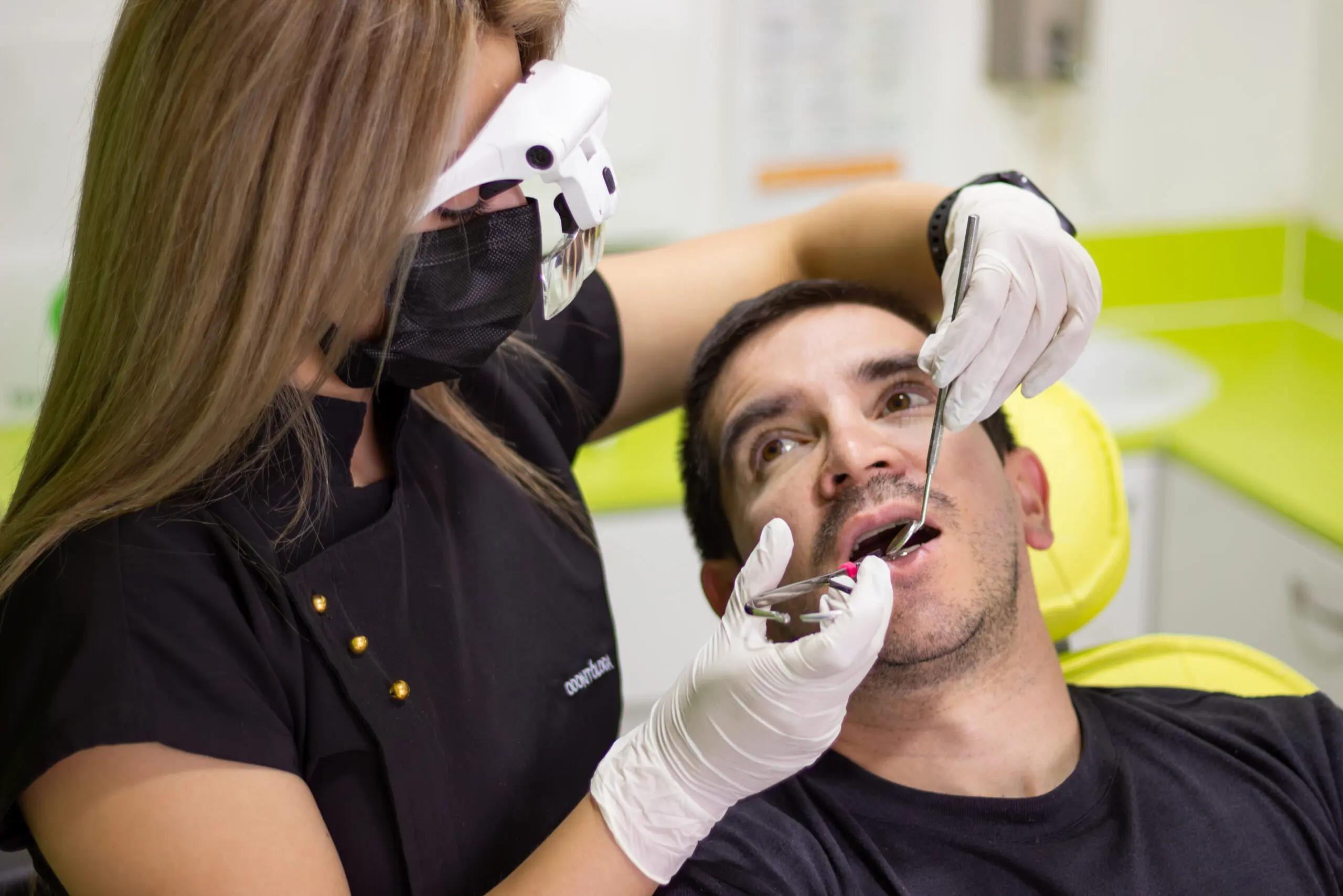 Female dentist wearing loupes and mask performing a dental examination on a male patient in a bright modern clinic, focusing on oral health and hygiene