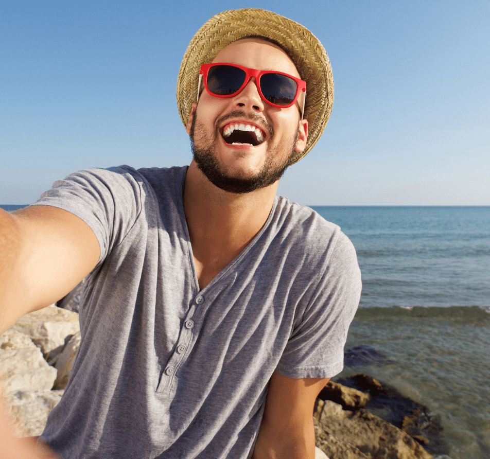 Adult man taking a selfie outdoors at the beach wearing a hat and sunglasses.