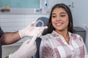 Delighted Hispanic female teenager with long dark hair in casual clothes smiling while looking at transparent vacuum formed retainer in hands of crop anonymous male dentist